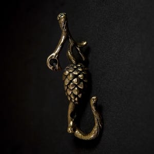 May include: A bronze-colored hook shaped like a pinecone and branch. The hook has a detailed, textured surface and a curved bottom. The design is nature-inspired, with a small, dark accent near the top.