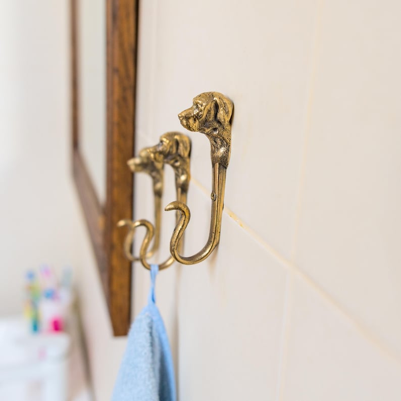 May include: Three gold-toned dog head hooks mounted on a white tiled wall. A blue towel hangs from one of the hooks.
