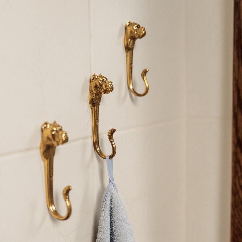 Op de afbeelding: Drie gouden haken in de vorm van hondenkoppen, gemonteerd op een witte betegelde muur. Een blauwe handdoek hangt aan een van de haken.