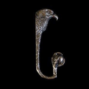 Op de afbeelding: Een zilverkleurige metalen haak in de vorm van een adelaar. De haak heeft een adelaarskop aan de bovenkant en een klauw die een bol vasthoudt aan de onderkant. De haak is ontworpen om items op te hangen en heeft een gedetailleerd, getextureerd oppervlak.