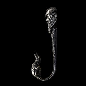 Könnte beinhalten: Ein silberfarbener Metallhaken mit detailliertem Design. Der Haken zeigt oben einen Wikingerhelm und ein Gesicht, unten einen Vogel. Der Haken ist gebogen und hat auf seiner gesamten Länge komplizierte Muster, vor einem schwarzen Hintergrund.