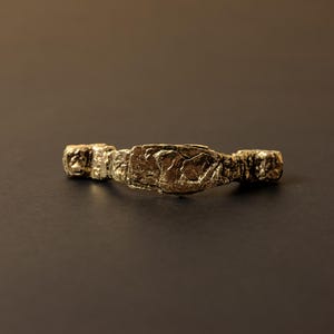 Puede incluir: Un brazalete dorado texturizado, con un centro rectangular y extremos redondeados. El brazalete tiene una apariencia rugosa y orgánica, lo que sugiere un diseño artesanal o vintage. El metal tiene un aspecto ligeramente deslustrado.