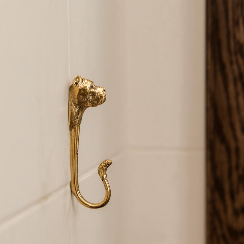 Op de afbeelding: Een gouden metalen haak in de vorm van een hondenkop. De haak is bevestigd aan een witte betegelde muur.