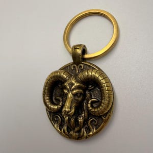 May include: A bronze-colored keychain featuring a detailed relief of a ram's head. The ram's head is centered on a circular pendant, with a textured surface and a gold-colored key ring attached. The keychain is made of metal and has an antique finish.