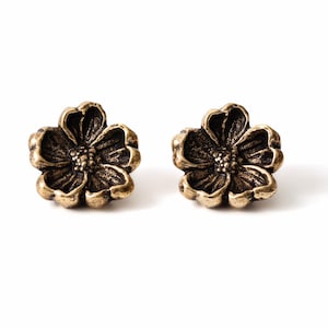 Peut inclure: Une paire de boucles d'oreilles à clous en forme de fleurs à cinq pétales. Les boucles d'oreilles sont fabriquées en métal bronze vieilli foncé avec une finition texturée et en creux qui souligne les nervures détaillées des pétales et le petit centre perlé.