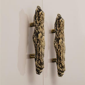 May include: Two gold-toned cabinet pulls shaped like rough-hewn pieces of wood. The pulls have a textured surface and are mounted on a white wall.