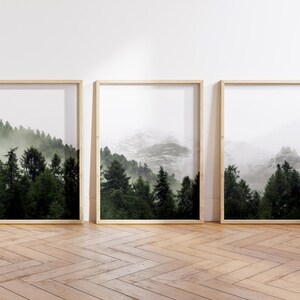May include: Three framed prints of a misty forest scene with mountains in the background. The prints are in wooden frames and are hanging on a white wall.