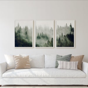 May include: A white sofa with three throw pillows sits in front of a white wall with three framed prints of a foggy forest scene. Two lamps with white lampshades are on end tables on either side of the sofa.