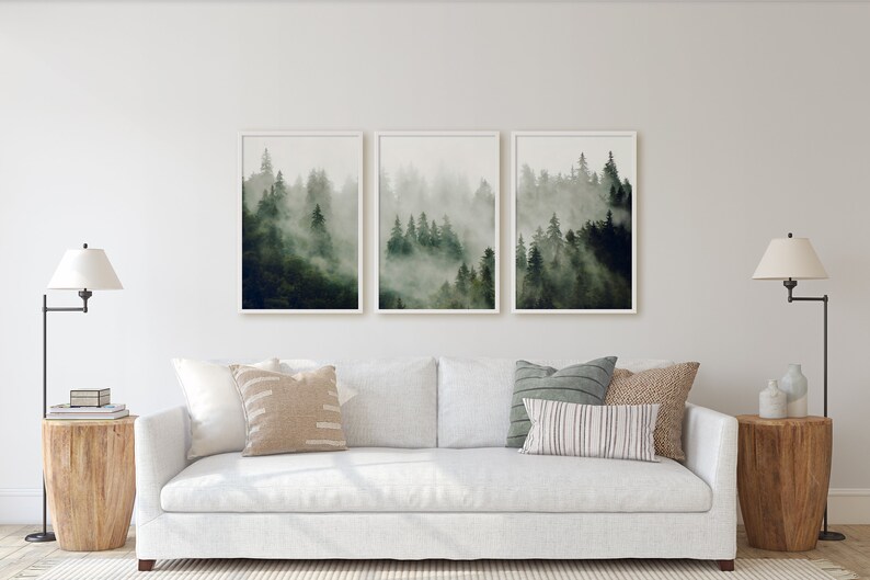 May include: A white sofa with three throw pillows sits in front of a white wall with three framed prints of a foggy forest scene. Two lamps with white lampshades are on end tables on either side of the sofa.