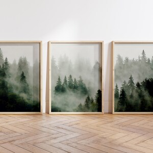 May include: Three framed prints of a misty forest scene with tall, dark green trees. The prints are arranged in a row on a light wood floor.