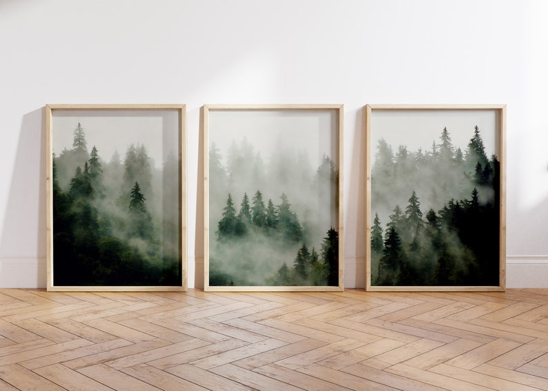 May include: Three framed prints of a misty forest scene with tall, dark green trees. The prints are arranged in a row on a light wood floor.