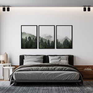 May include: A gray upholstered bed with a black headboard and frame, set against a white wall with three framed prints of a foggy forest scene. The bed is dressed with gray linens and pillows. A small white side table with a lamp and vase sits next to the bed.