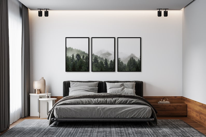 May include: A gray upholstered bed with a black headboard and frame, set against a white wall with three framed prints of a foggy forest scene. The bed is dressed with gray linens and pillows. A small white side table with a lamp and vase sits next to the bed.