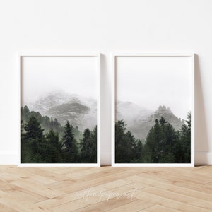 May include: Two framed prints of a misty mountain landscape with a forest in the foreground. The prints are in white frames and are hung on a white wall.