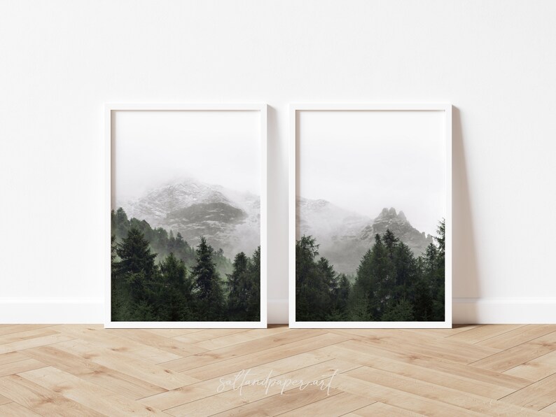 May include: Two framed prints of a misty mountain landscape with a forest in the foreground. The prints are in white frames and are hung on a white wall.