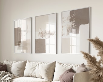 May include: Three framed abstract art prints with brown and beige brushstrokes on a white background. The prints are hanging on a white wall above a beige couch with pillows.