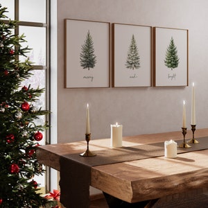 Pode incluir: Tr&ecirc;s pinturas de aquarela emolduradas de pinheiros com as palavras "merry", "and" e "bright" abaixo de cada &aacute;rvore. As pinturas est&atilde;o penduradas em uma parede branca sobre uma mesa de madeira com um corredor marrom e velas acesas.