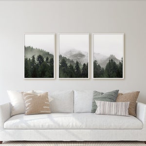 May include: Three framed prints of a misty forest scene with a mountain range in the background. The prints are hung on a white wall above a white sofa with pillows.