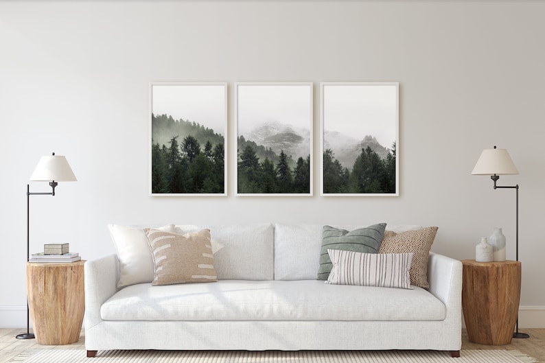 May include: Three framed prints of a misty forest scene with a mountain range in the background. The prints are hung on a white wall above a white sofa with pillows.