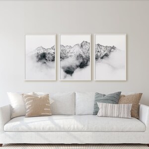 May include: A white sofa with three pillows, two lamps, and a set of three framed black and white photographs of mountains with clouds above them.