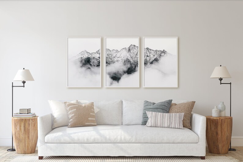 May include: A white sofa with three pillows, two lamps, and a set of three framed black and white photographs of mountains with clouds above them.