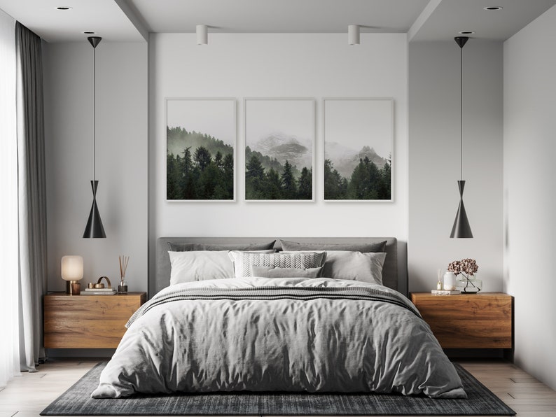 May include: A bedroom interior with a gray bed, two wooden nightstands, and three framed landscape photographs of a misty forest. The room has a modern minimalist design with white walls and black pendant lights.