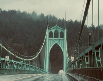Könnte beinhalten: Eine türkisgrüne Hängebrücke mit einem gotischen Torbogen über einer nassen Straße. Die Brücke hat ein Tempolimit von 48 km/h. Ein Auto fährt auf den Torbogen zu. Im Hintergrund befindet sich ein dichter Wald unter einem bewölkten Himmel.