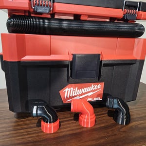 Puede incluir: Una caja de herramientas Milwaukee Fuel roja y negra con una manguera flexible negra acoplada. Frente a la caja hay tres adaptadores de manguera de aspiradora en forma de L, dos negros y uno rojo, todos con un diseño liso y curvo.