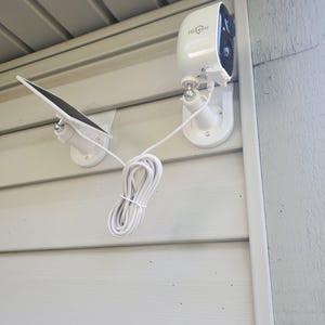 Puede incluir: Sistema de cámara de seguridad blanco con panel solar, montado en una pared exterior gris claro. La cámara tiene la marca "HiSeeu" impresa. Un cable blanco conecta la cámara al panel solar.