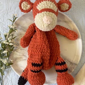 May include: A handmade orange and cream-colored crochet tiger plush toy. The tiger has black stripes on its legs and tail, with a pink nose and cream-colored accents on its face and ears. The plush is sitting on a white plate.