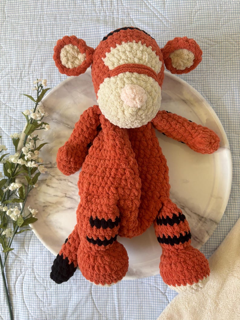 May include: A handmade orange and cream-colored crochet tiger plush toy. The tiger has black stripes on its legs and tail, with a pink nose and cream-colored accents on its face and ears. The plush is sitting on a white plate.