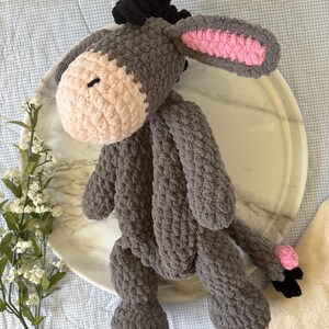 May include: A handmade crochet donkey plushie in shades of gray, with a light pink muzzle and inner ears. The donkey has black hair and a small black tail. The plushie is sitting on a marble tray.