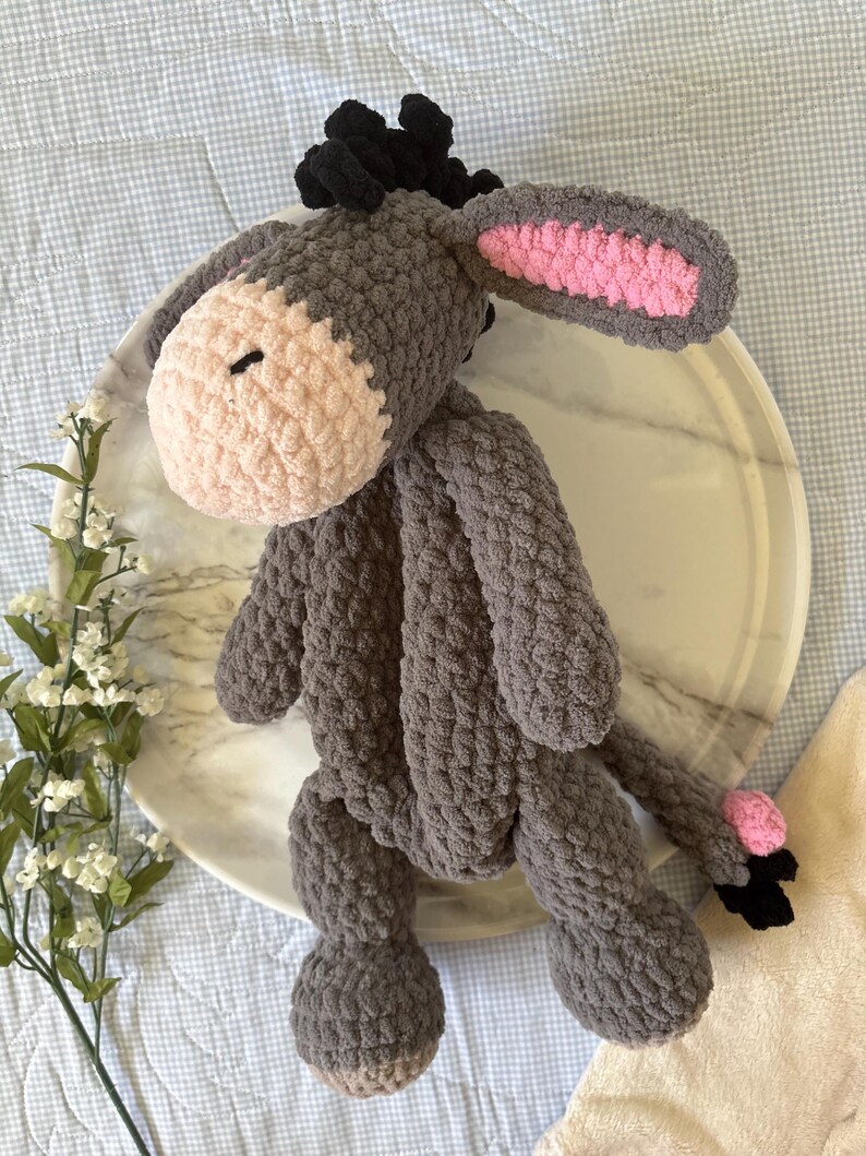 May include: A handmade crochet donkey plushie in shades of gray, with a light pink muzzle and inner ears. The donkey has black hair and a small black tail. The plushie is sitting on a marble tray.