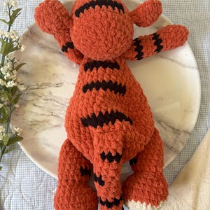 May include: A handmade orange and black crochet tiger toy with white accents. The plush tiger has black stripes and is lying on a white marble plate. The toy has outstretched arms and a tail.