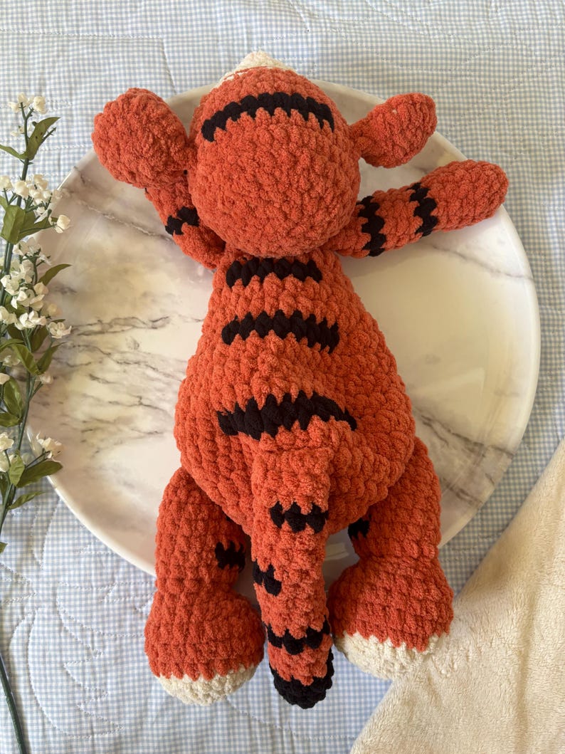 May include: A handmade orange and black crochet tiger toy with white accents. The plush tiger has black stripes and is lying on a white marble plate. The toy has outstretched arms and a tail.