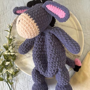May include: A handmade plush donkey toy in shades of purple, pink, and black. The crochet donkey has a light pink muzzle, inner ears, and a small pink tail. The donkey is sitting on a white marble plate.