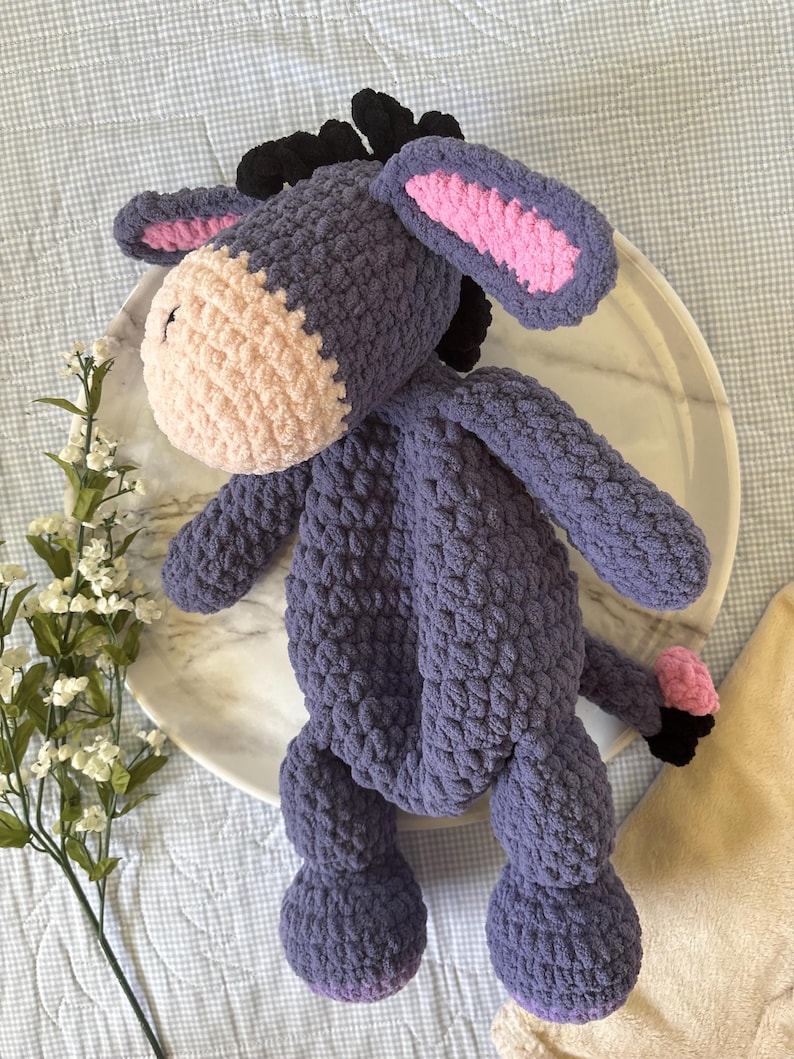 May include: A handmade plush donkey toy in shades of purple, pink, and black. The crochet donkey has a light pink muzzle, inner ears, and a small pink tail. The donkey is sitting on a white marble plate.