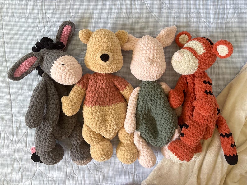 May include: Four handmade crochet plush toys: a gray donkey with pink ears, a yellow bear with an orange shirt, a pink pig with a green dress, and an orange tiger with black stripes. The toys are arranged on a light blue surface.