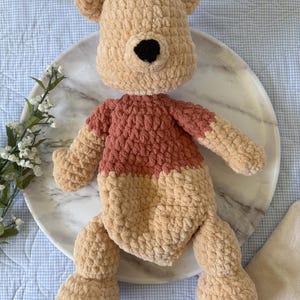 May include: A handmade crochet plush toy in the shape of a bear. The bear has a tan head and limbs, a rust-colored shirt, and a black nose. The toy is sitting on a marble plate.