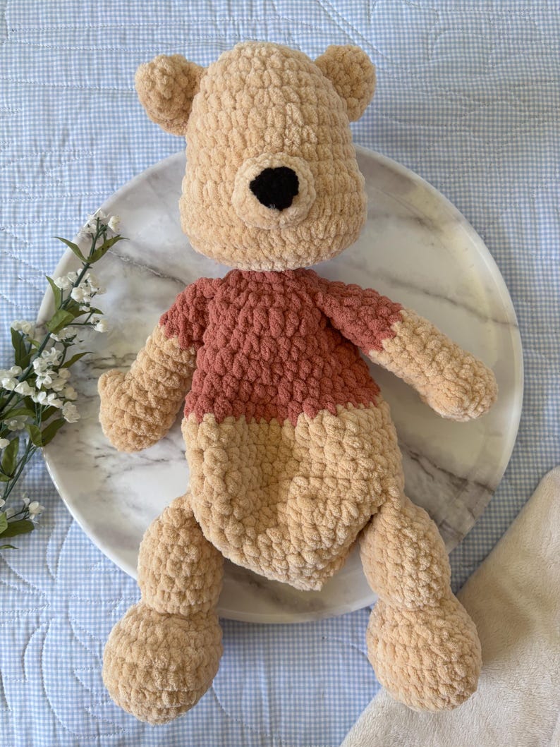 May include: A handmade crochet plush toy in the shape of a bear. The bear has a tan head and limbs, a rust-colored shirt, and a black nose. The toy is sitting on a marble plate.