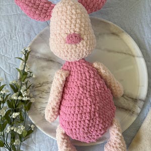 May include: A handmade crochet pig plush toy. The pig has a light pink body, arms, and legs, with a darker pink belly, ears, and snout. The toy is sitting on a white marble plate.