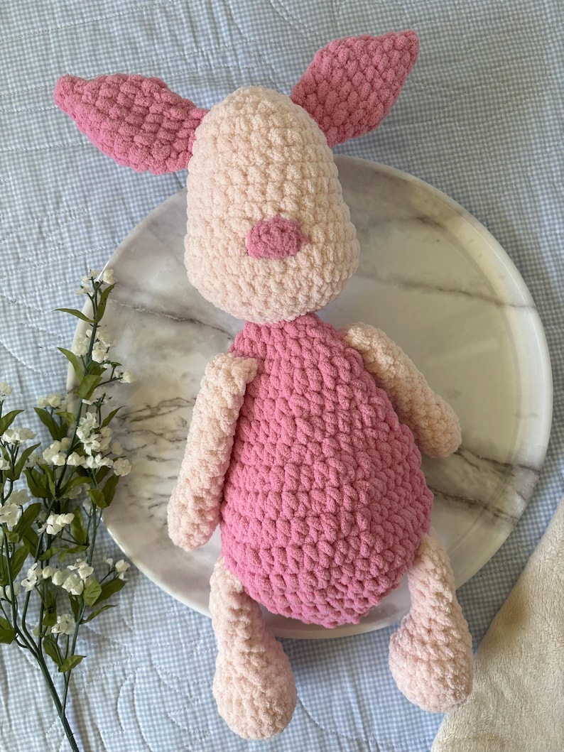 May include: A handmade crochet pig plush toy. The pig has a light pink body, arms, and legs, with a darker pink belly, ears, and snout. The toy is sitting on a white marble plate.