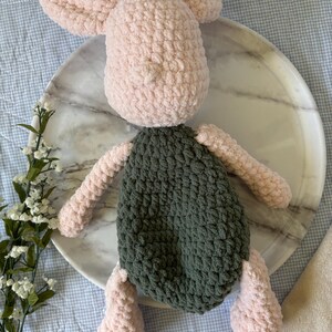 May include: A handmade crochet pig plush toy. The pig has a light pink head, ears, arms, and legs, and a dark green body. The plush is sitting on a marble tray.