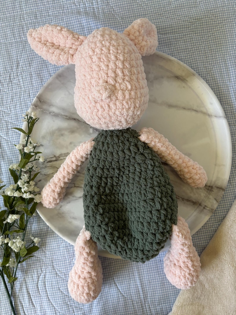 May include: A handmade crochet pig plush toy. The pig has a light pink head, ears, arms, and legs, and a dark green body. The plush is sitting on a marble tray.