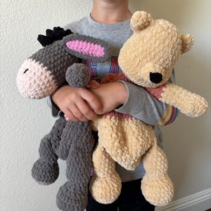 May include: Two handmade crochet stuffed animals: a gray donkey with pink ears and a tan bear with a red scarf. The donkey has a black mane and the bear has a black nose. Both are soft and plush.