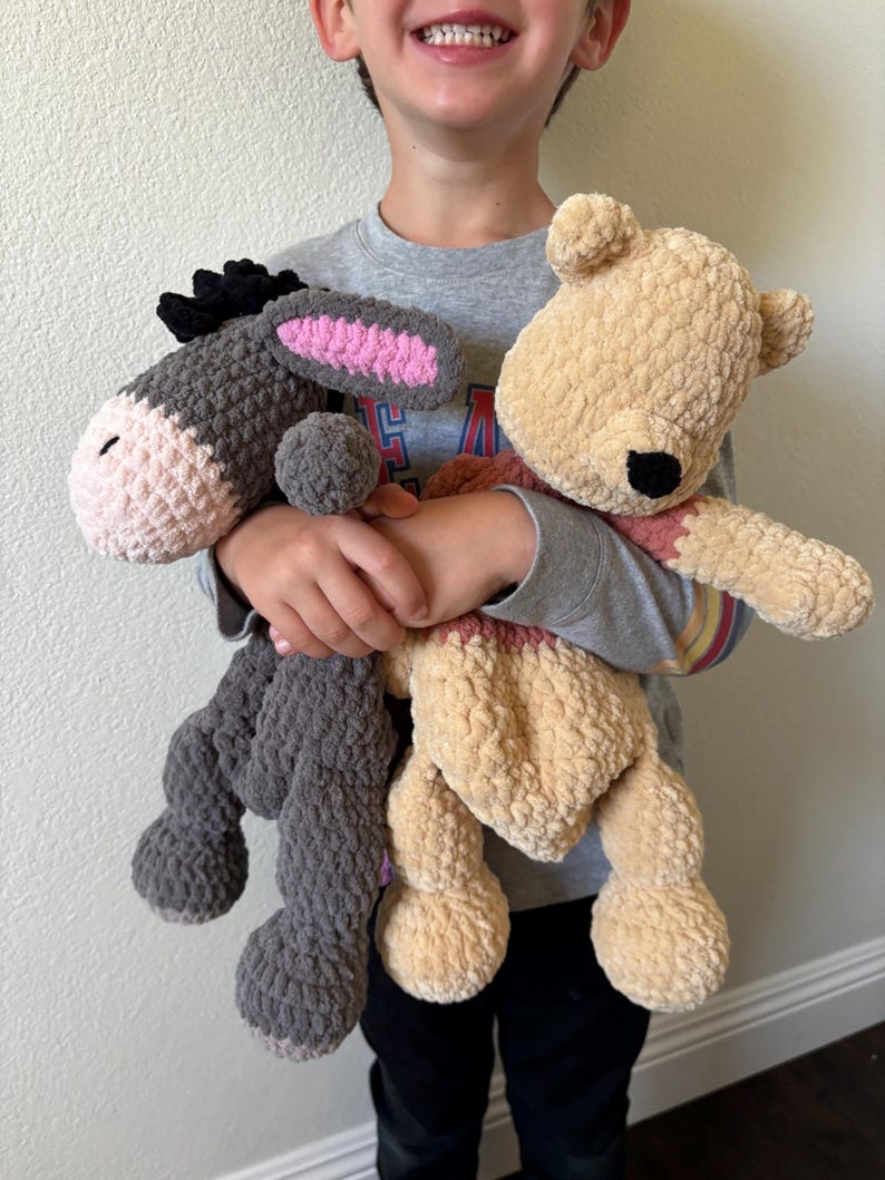 May include: Two handmade crochet stuffed animals: a gray donkey with pink ears and a tan bear with a red scarf. The donkey has a black mane and the bear has a black nose. Both are soft and plush.