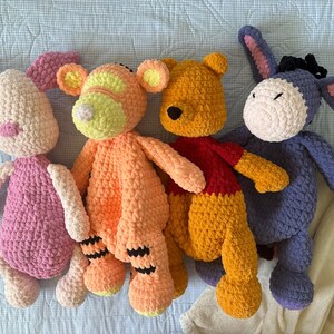 May include: Four handmade crochet plush toys: a pink Piglet, an orange Tigger, a yellow Winnie the Pooh wearing a red shirt, and a purple Eeyore. Each plush has a unique design and color scheme, perfect for children.