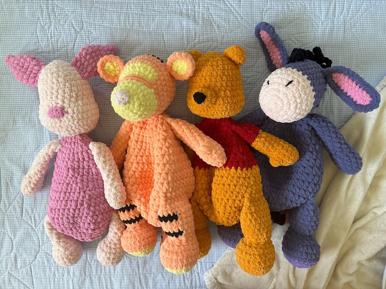 May include: Four handmade crochet plush toys: a pink Piglet, an orange Tigger, a yellow Winnie the Pooh wearing a red shirt, and a purple Eeyore. Each plush has a unique design and color scheme, perfect for children.