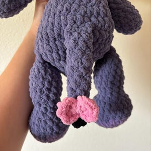 May include: A handmade, plush toy in a soft, textured purple yarn. The toy has large, rounded legs and a body with a textured surface. The feet are pink with black accents. The toy is designed to be cuddly.