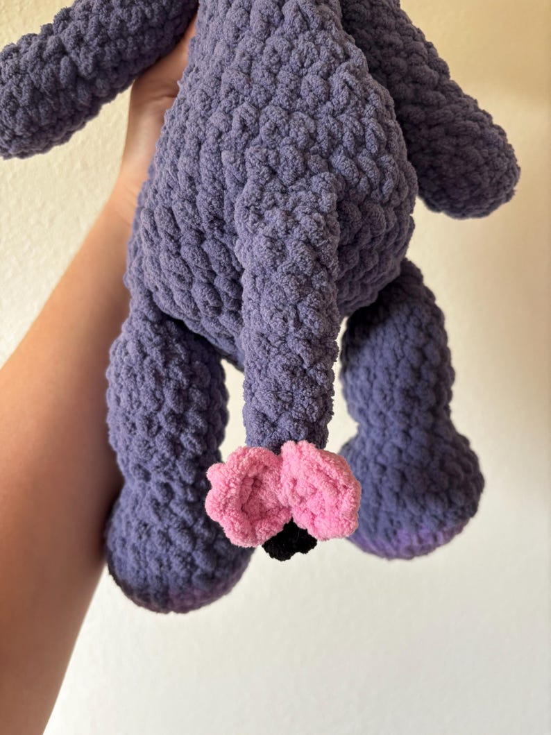 May include: A handmade, plush toy in a soft, textured purple yarn. The toy has large, rounded legs and a body with a textured surface. The feet are pink with black accents. The toy is designed to be cuddly.